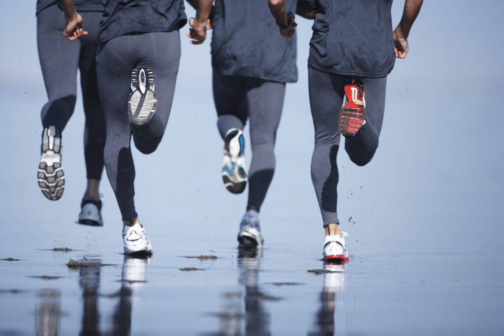 Four men jogging on beach, low section, rear view